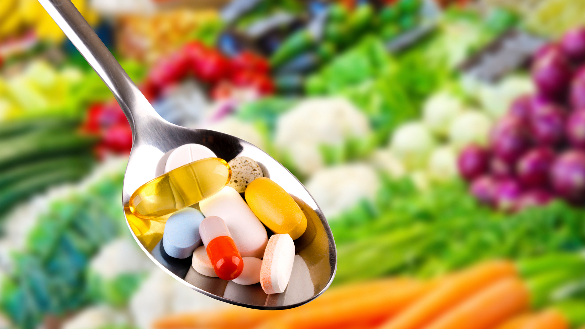 Imagem com uma colher segurando diversas capsulas coloridas e ao fundo diversos legumes coloridos