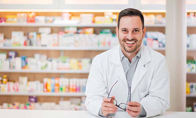 farmacêutico sorrindo, com um jaleco branco, segurando um óculos, atrás é possível ver diversos medicamentos