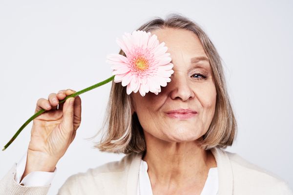 Mulher mais velha sorrindo, com cabelos curtos e loiros escuros, segurando uma flor verde e rosa em cima de um dos olhos