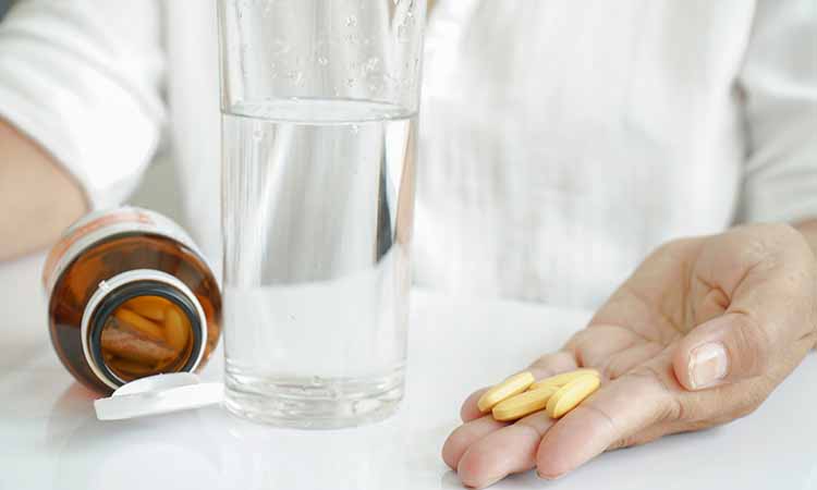 mulher segurando capsulas amarelas na mãos, um copo de água e um pote de remédio aberto com medicamento dentro