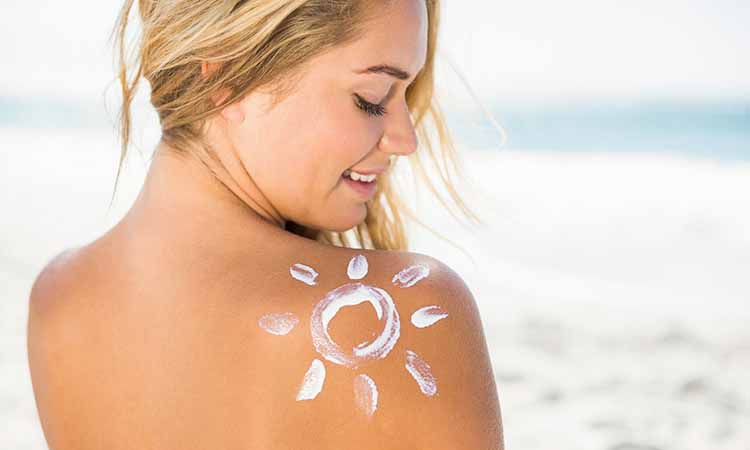 Mulher loira de costas sorrindo em uma praia com um desenho de sol feito pelo protetor solar