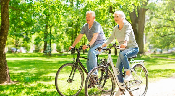 Um casal de idosos andando de bicicleta em uma parque com arvores ao redor